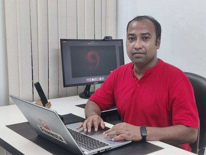Photo of Ashique Rahman, a Bangladeshi man sitting at a desk with a laptop in front of him, wearing a red v-neck shirt.