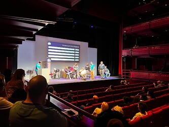 People sat in a conference hall. There are several people on stage playing a game of Pointless