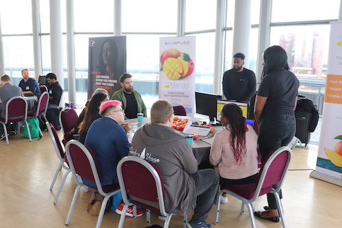 A group of people sat around a circular table at a conference. There are two people stood up running an activity with them.
