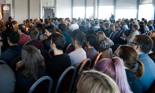 A crowd of people sat in a conference room listening to a speaker on stage. The person on stage is just out of shot on the left.