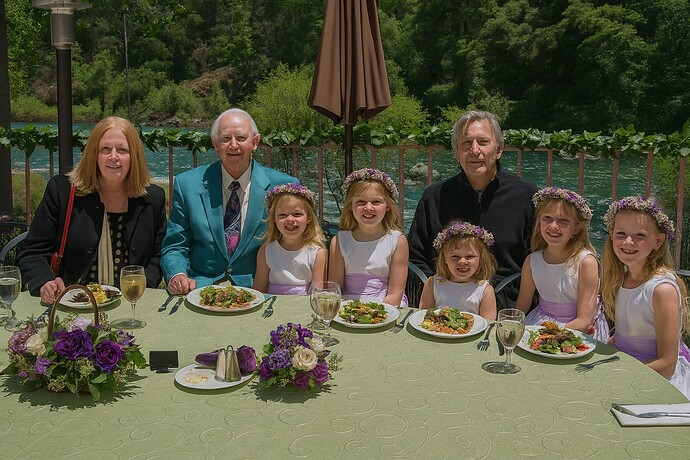 A group of people, including two men, one woman, and five young girls in matching white dresses with flower crowns, are seated at a round outdoor table set with food and flowers, with a scenic river and greenery in the background. (Captioned by AI)